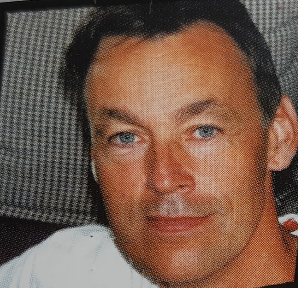 Close-up portrait of a middle-aged man with blue eyes, short dark hair, and light stubble.