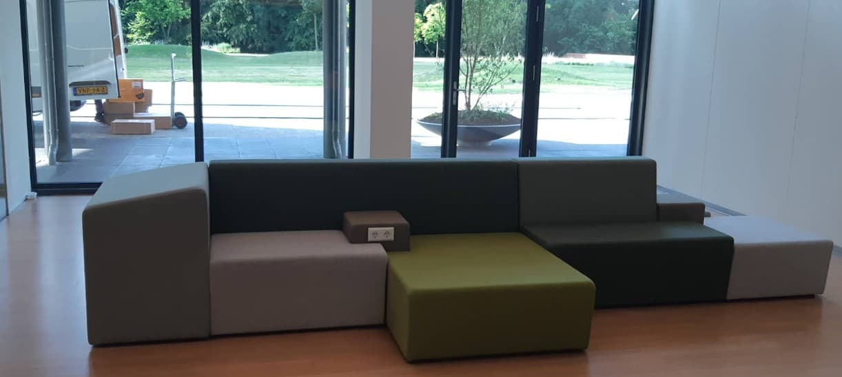 Modulaire bank in aardetinten met een geïntegreerd stopcontact, geplaatst in een lichte, moderne ruimte met grote raampartijen.